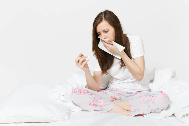 Chory zmęczony smutny Kobieta w piżamie, siedząc na łóżku, trzymając kliniczne termometr z wysoką gorączką temperatury izolowana na białym tle. Kobieta czuje się źle, dmuchanie jej nos do serwetka. - Zdjęcie, obraz