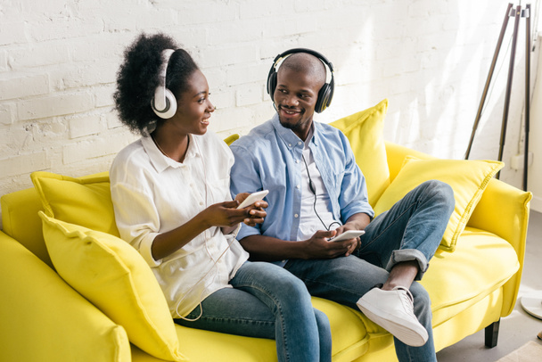 african american listening music in headphones with smartphones while resting on sofa at home - Foto, Bild