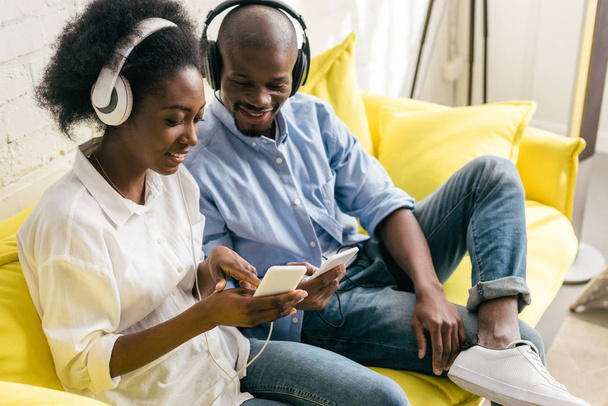 smiling african american listening music in headphones and using smartphones while resting on sofa at home - Foto, Imagen