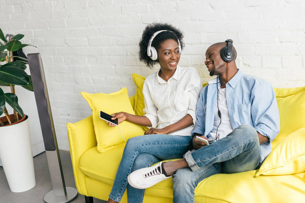 sorridente musica d'ascolto africana americana in cuffia con smartphone mentre si riposa sul divano di casa
 - Foto, immagini