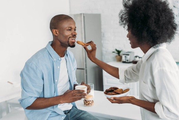 mujer afroamericana alimentando marido con galletas en la mañana en la cocina en casa
 - Foto, imagen