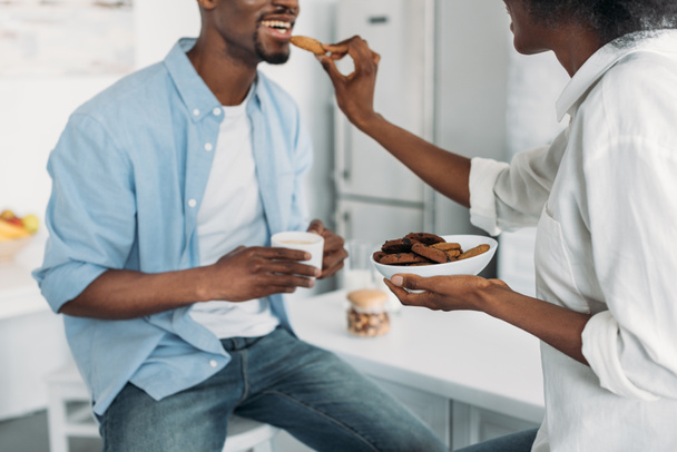 partial view of african american woman feeding husband with cookies in morning in kitchen at home - Foto, Bild