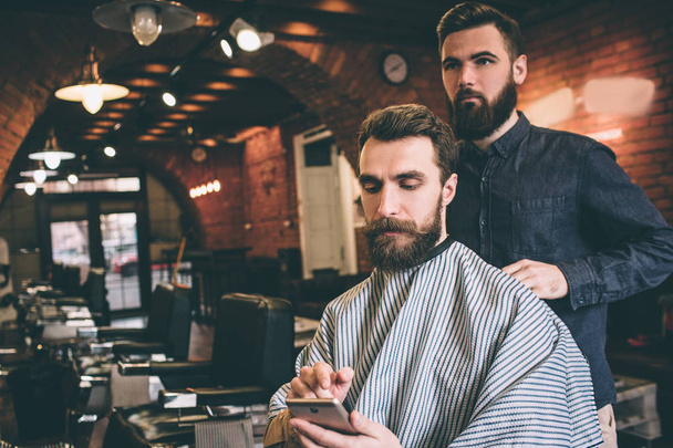 Bearded guy is sitting in chair and getting ready for procedure. He is looking at the phone. He is very concentrated. Hairdresser is ready to start procedure. - Photo, Image