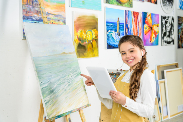 smiling child standing with tablet in workshop of art school and pointing on canvas - Photo, Image