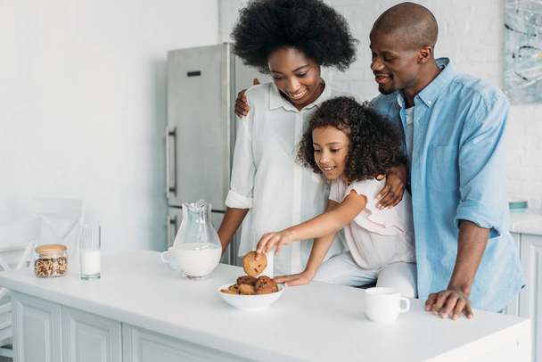 african american kid taking cookie while parents standing near by in kitchen at home - Zdjęcie, obraz