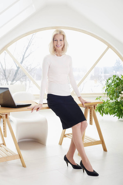 Full length shot of beautiful businesswoman with toothy smile standing at office desk and looking at camera. - Valokuva, kuva
