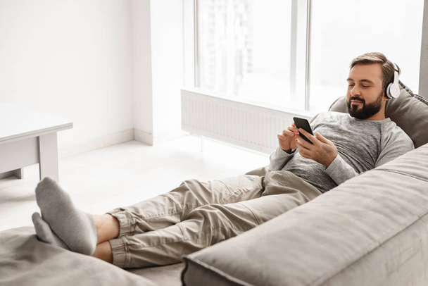Young guy 30s in basic clothing lying on sofa at home, and liste - Foto, Imagem