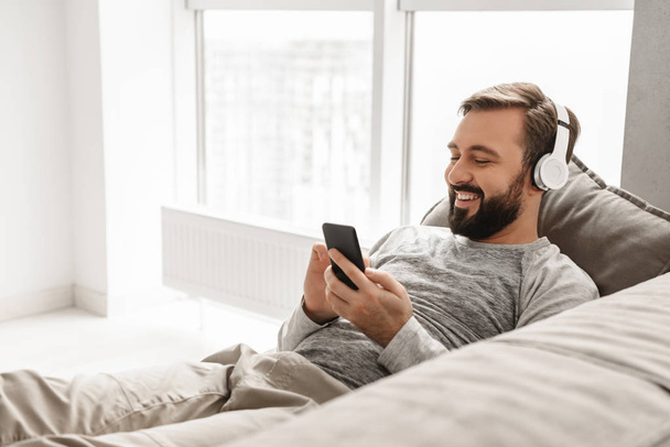 Bearded man 30s in basic clothing lying on sofa at home, and lis - 写真・画像