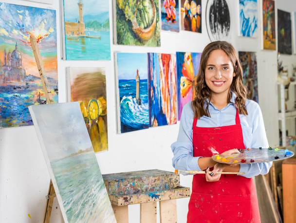 atractiva artista femenina sosteniendo pincel de pintura y paleta cerca de lienzo en taller
 - Foto, imagen