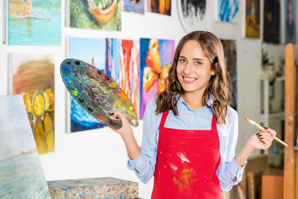 sonriente hermosa artista femenina sosteniendo pincel de pintura y paleta en el taller
 - Foto, Imagen