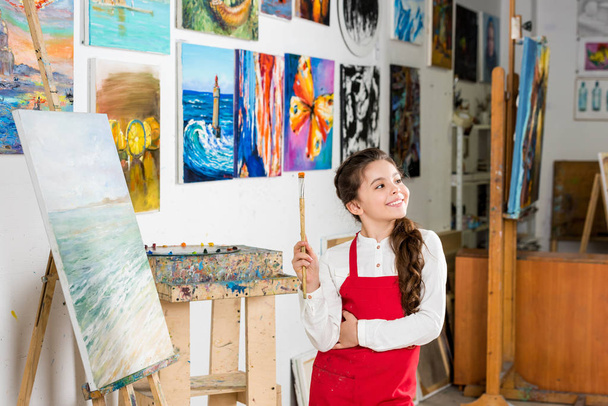 Kind hält Malpinsel in der Werkstatt der Kunstschule und schaut weg - Foto, Bild