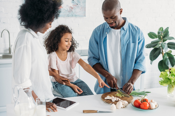african american man cutting fresh vegetables for breakfast with family standing near by in kitchen at home - Foto, afbeelding