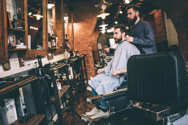 A picture from a distance. Guy is sitting in a chair and getting ready for cutting beard. His hairstylist is ready to start to work. - Foto, immagini