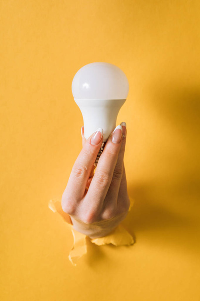 Hand holding a light bulb breaking through the hole in yellow background - Foto, Imagen