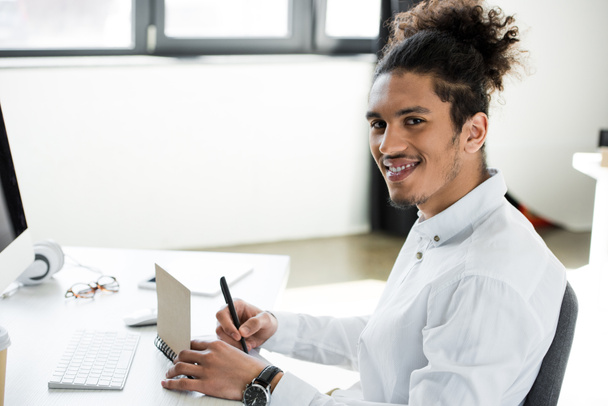 handsome young african american businessman smiling at camera while taking notes at workplace - 写真・画像