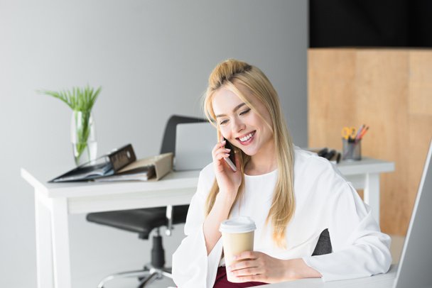lachende jonge zakenvrouw holding wegwerp koffiekopje en praten door smartphone in kantoor - Foto, afbeelding