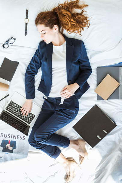 Top view of businesswoman in suit typing on laptop while lying in bed among folders and documents - Photo, Image