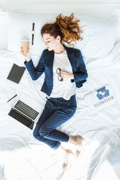 Pohled shora podnikatelka v obleku drží šálek kávy a spí v posteli mezi složek a dokumentů - Fotografie, Obrázek