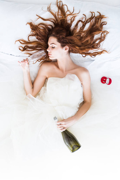 Κάτοψη του νύφη φορώντας λευκό φόρεμα ξαπλωμένη στο κρεβάτι και πίνοντας σαμπάνια - Φωτογραφία, εικόνα