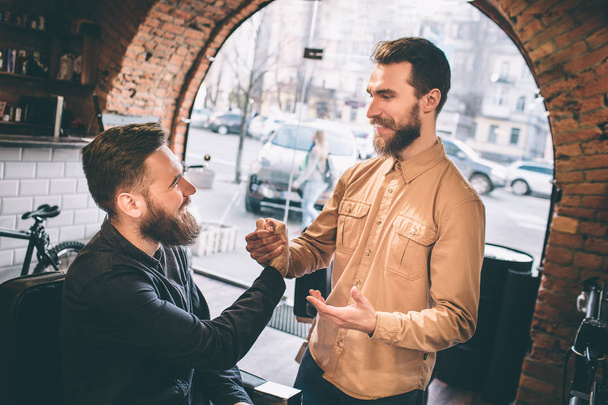 Two friends are in a barbershop. They are shaking each others hands. Guys are smiling to each other. - Foto, afbeelding