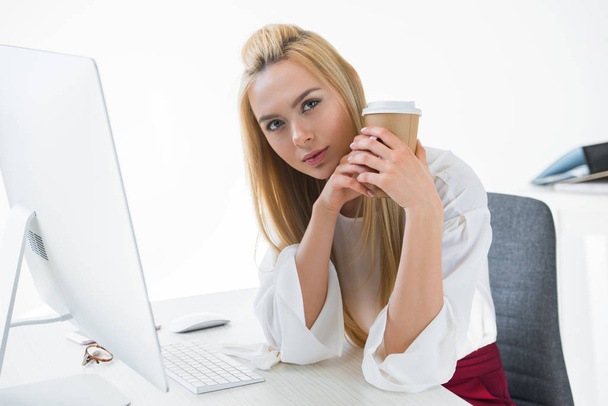 beautiful young businesswoman holding disposable coffee cup and looking at camera in office - Foto, Imagen