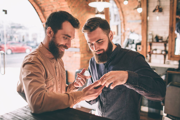 Two happy friends are in a barbershop. They are standing together and looking at the phone. One of them is showing something on the phone to the other guy. - Foto, Imagen