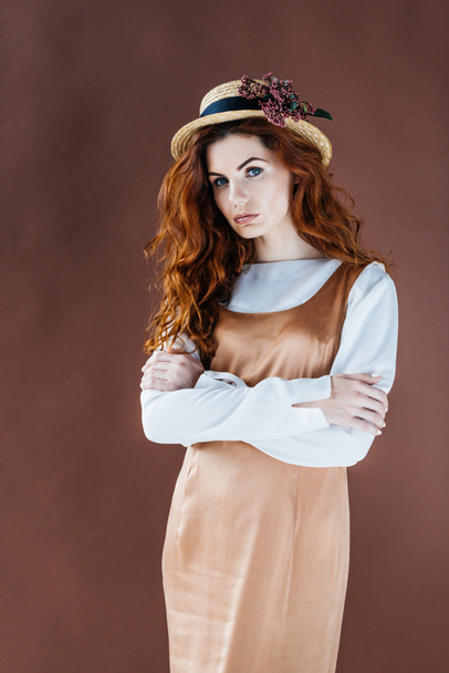 Woman with long red hair in beige dress and straw hat isolated on brown background - Photo, image