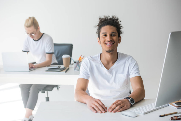 young african american man smiling at camera while working in office with female colleague behind - Foto, Imagen