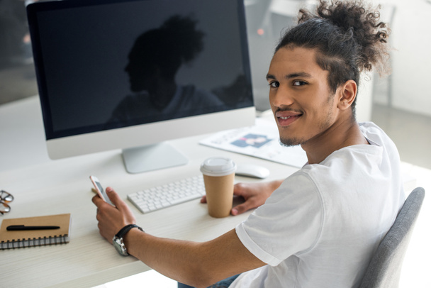 joven afroamericano hombre con taza de papel y teléfono inteligente sonriendo a la cámara en el lugar de trabajo
 - Foto, Imagen