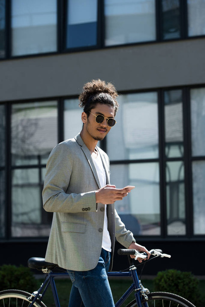 jovem homem americano africano olhando para a câmera enquanto está de pé com bicicleta e usando smartphone
 - Foto, Imagem