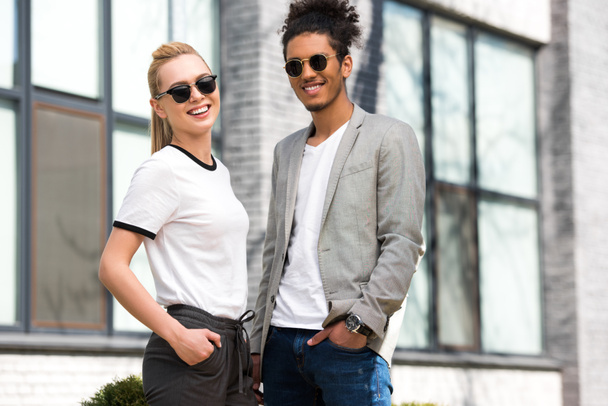 retrato de belo casal multiétnico elegante em óculos de sol de pé com as mãos em bolsos e sorrindo para a câmera
 - Foto, Imagem