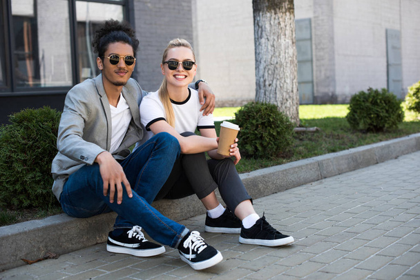 happy young multiethnic couple smiling at camera while sitting on street with coffee to go  - 写真・画像