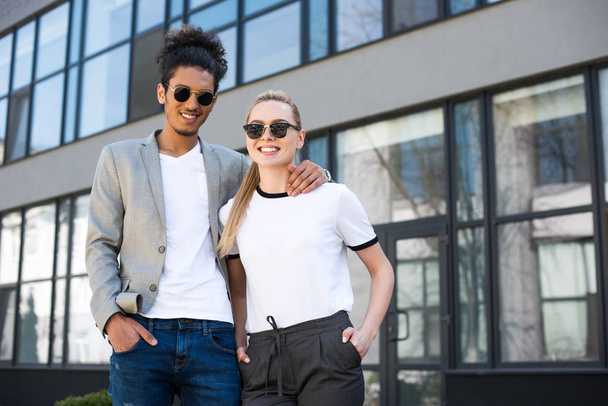 young multiethnic couple in sunglasses standing together and smiling at camera on street - Photo, image
