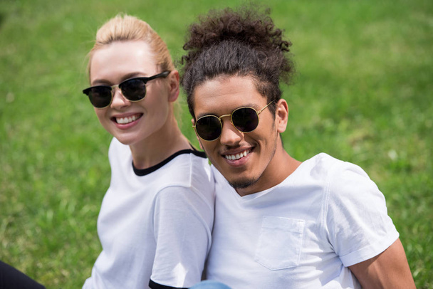 portrait of beautiful young multiethnic couple in sunglasses sitting together on grass and smiling at camera - Fotó, kép