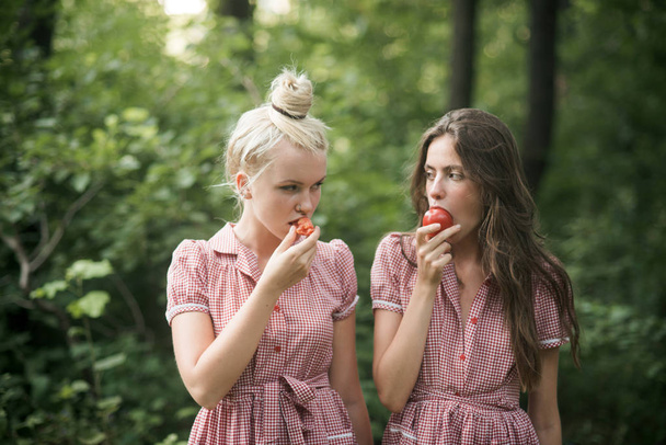 Camping in woods. Beautiful young girls wandering in forest. Two sisters in vintage dresses eating juicy tomatoes. Vitamins, nutrition and healthy diet concept. - Фото, изображение