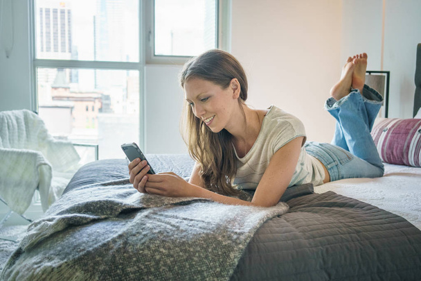 Glimlachend jonge vrouw messaging op mobiele telefoon in bed - Foto, afbeelding