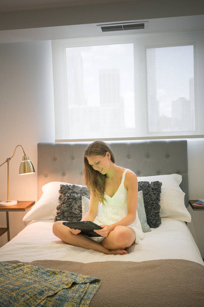 Gelukkig jonge vrouw in nachtkleding met behulp van digitale tablet in bed - Foto, afbeelding