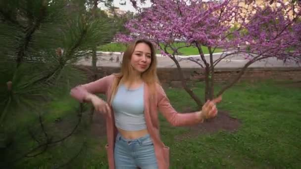Jocund dziewczyna taniec w pobliżu kwitnących drzew w parku i dmuchanie pocałunek. - Materiał filmowy, wideo