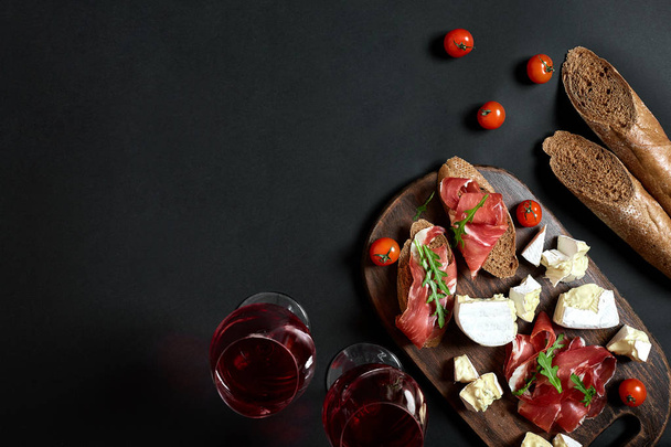 Prosciutto, salami, baguette slices, tomatoes and nutson rustic wooden board, two glasses of red wine over black background - Fotó, kép