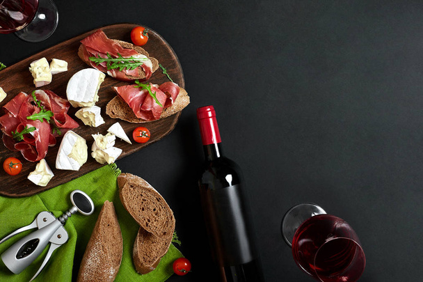 Herkullinen alkupala viinille kinkku, juusto, patonki viipaleet, tomaatit, tarjoillaan puulaudalla ja lasi punaviinillä mustalla pinnalla
 - Valokuva, kuva