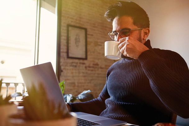 Cute handsome bearded man using mobile laptop computer while drinking coffee at living room at home. Man using electronic gadget. Blurred background. - 写真・画像