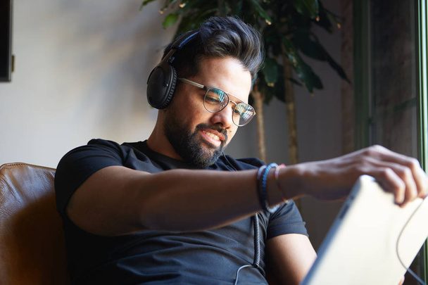 Handsome bearded man in headphones listening to music on electronic tablet at home. Relaxing and rest time concept. Blurred background. - Fotografie, Obrázek