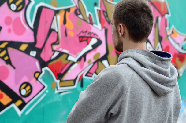 Egy fiatal graffiti művész egy szürke kapucnis úgy néz ki a fal a graffiti, rózsaszín és zöld színben a falra, az esős időjárás. Utcai művészeti koncepció - Fotó, kép