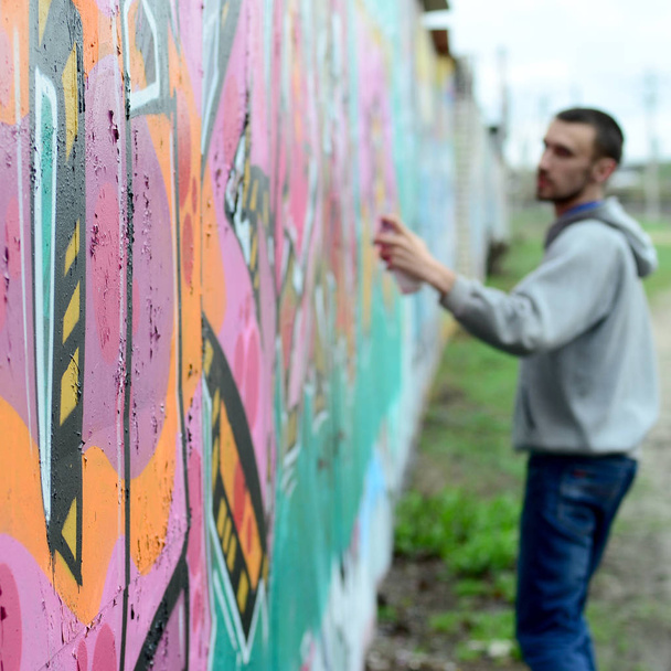 Young guy in a gray hoodie paints graffiti in pink and green colors on a wall in rainy weather Focus on the fragment of wall and blurred artist - Zdjęcie, obraz