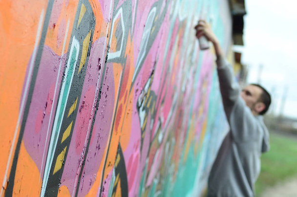 Young guy in a gray hoodie paints graffiti in pink and green colors on a wall in rainy weather Focus on the fragment of wall and blurred artist - Photo, Image