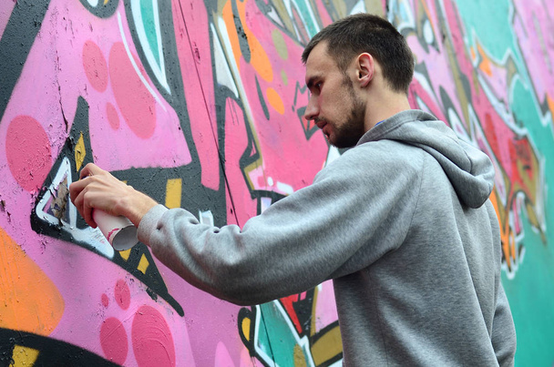 Ένας νεαρός άντρας σε ένα γκρι φούτερ με κουκούλα ζωγραφίζει γκράφιτι σε ροζ και πράσινο c - Φωτογραφία, εικόνα