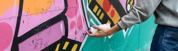 Hand of a young guy in a gray hoodie paints graffiti in pink and green colors on a wall in rainy weather - Foto, Imagen
