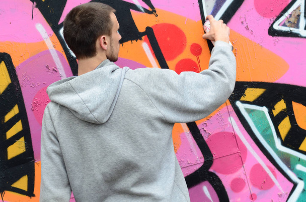 Mladý muž v šedém hoodie maluje graffiti v růžové a zelené barvy na zeď v deštivém počasí - Fotografie, Obrázek