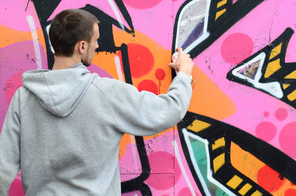 A young guy in a gray hoodie paints graffiti in pink and green colors on a wall in rainy weather - Photo, Image