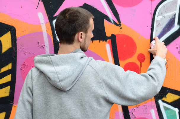 Gri kapüşonlu genç bir adam grafiti bir duvar pembe ve yeşil renklerde yağmurlu havalarda boyar. - Fotoğraf, Görsel